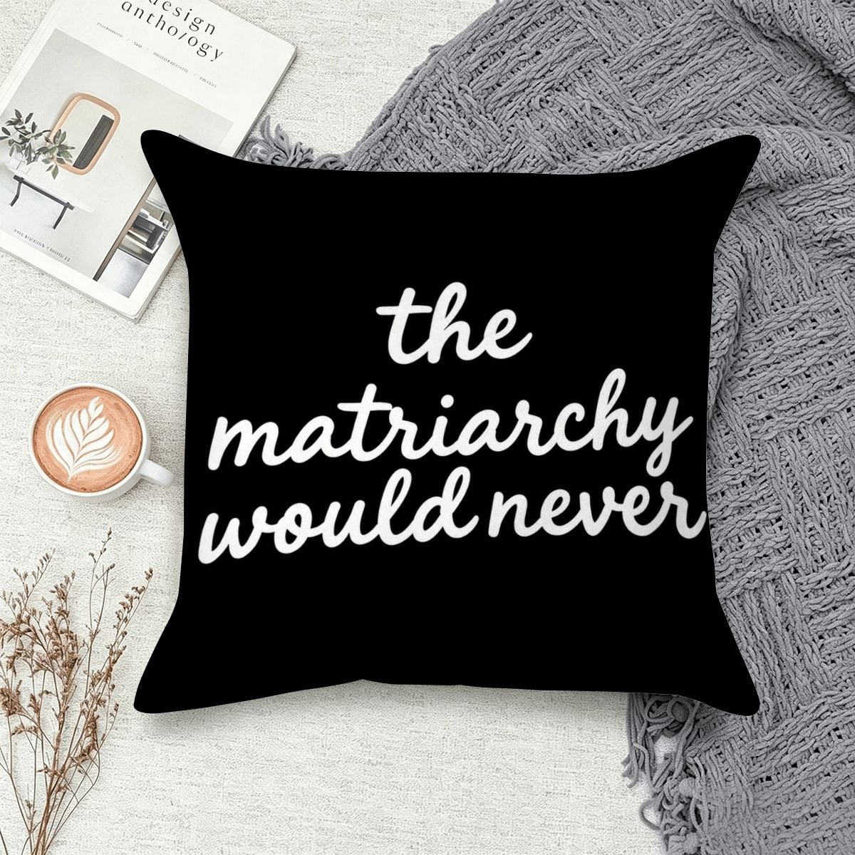 Black decorative throw pillow with white text reading 'the matriarchy would never' on a cozy surface.